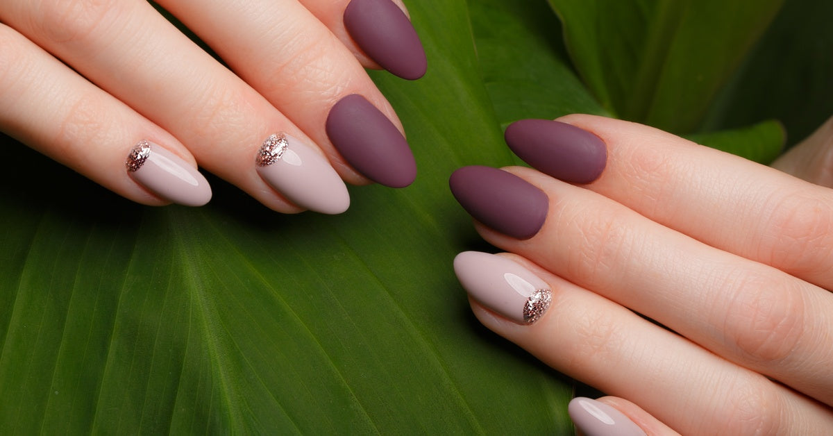A close-up of a neat acrylic purple matte manicure on a female's hands on a background of green leaves.